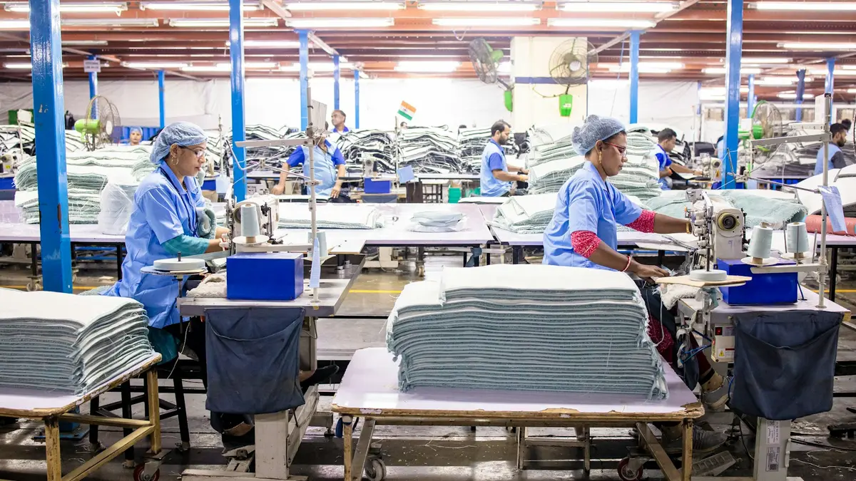 Workers at sewing machines in a textile factory, surrounded by neat stacks of fabric.