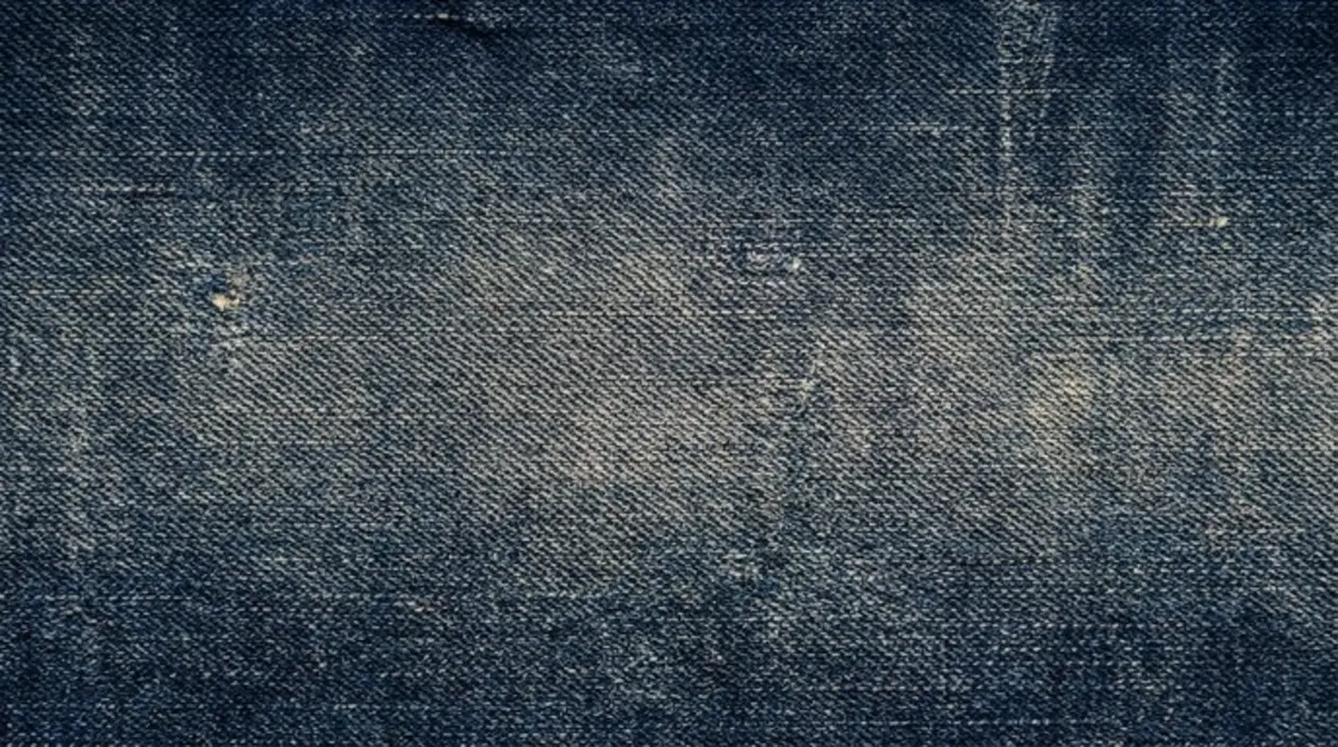 Close-up of blue denim fabric with visible slub texture, diagonal twill lines, faded patches, and irregular woven areas.