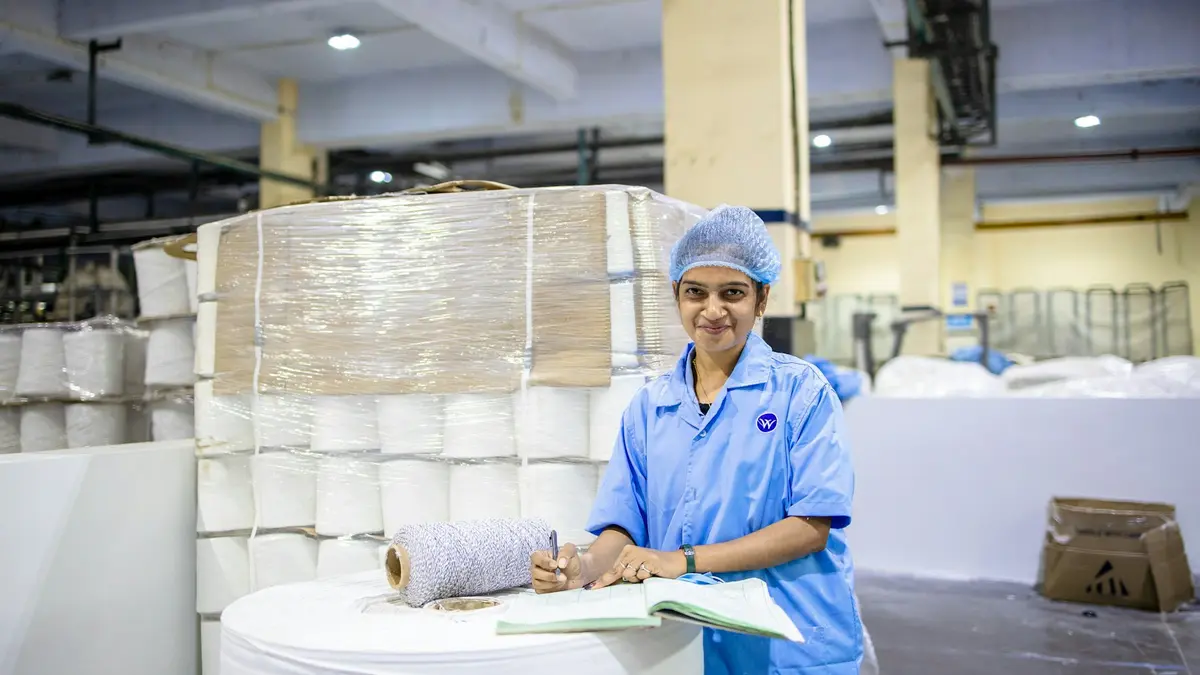 A woman in a blue uniform smiles while writing in a notebook beside large rolls of fabric.