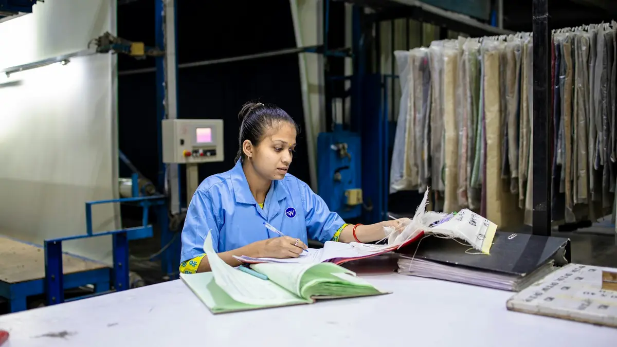 A woman in a blue uniform reviews documents at a textile facility surrounded by fabric samples.