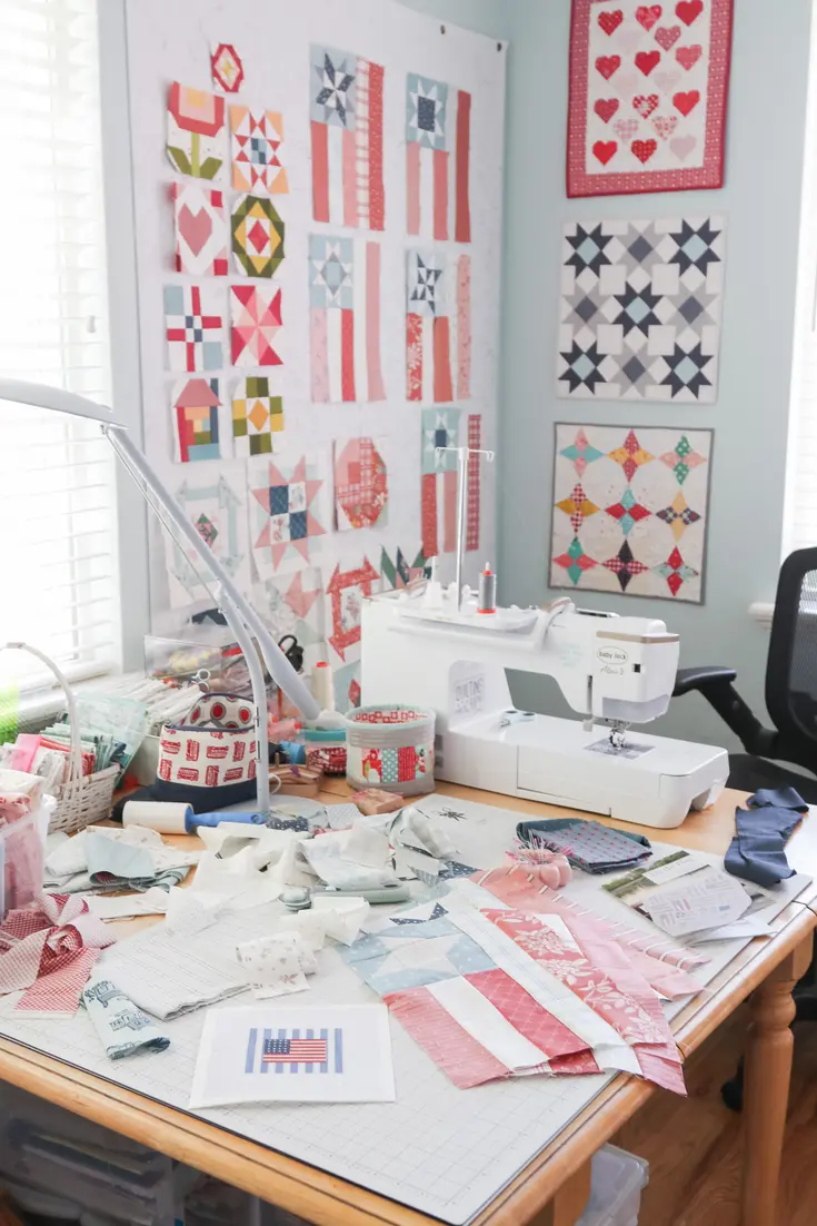 Sewing workspace with a machine, fabric swatches, garment pieces, and patchwork quilt designs displayed on the wall.